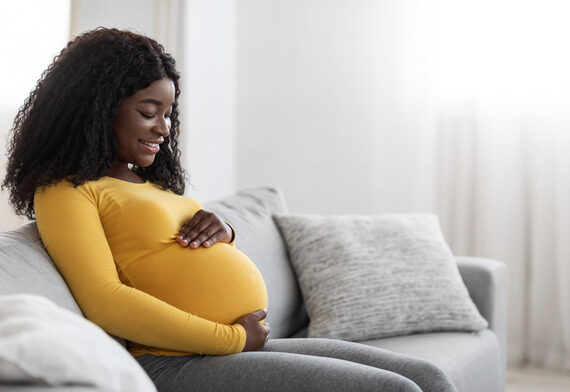 Joyful black pregnant woman hugging her big tummy, communicating with her baby, sitting on sofa at living room. Happy african american expecting lady spending time at home, copy space